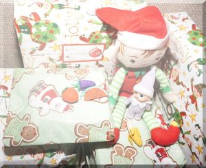 A Christmas elf sitting with a baby elf amongst a pile of presents and enjoying the scrunch of wrapping paper