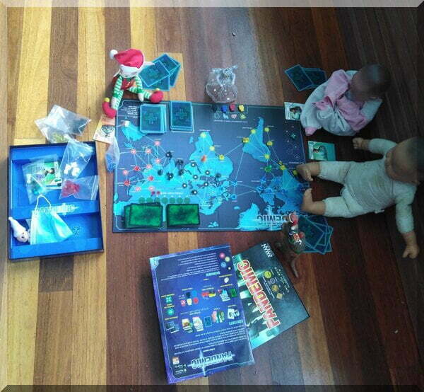 An angel and a Christmas elf called Tinkles playing the pandemic board game with some dolls on a wooden floor