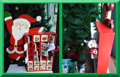A wooden Santa advent calendar with a baby CHristmas elf and reindeer puppet poking out of some doors