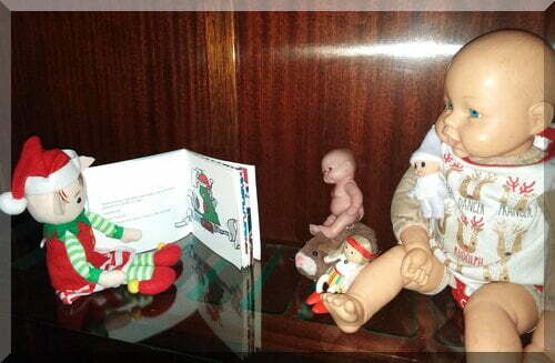 A Christmas elf sitting on a shelf reading a Christmas book to some dolls