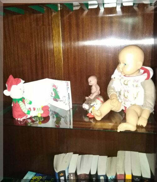 A Christmas elf sitting on a shelf reading a Christmas book to some dolls