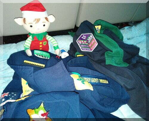 CHristmas elf sewing a badge onto a blue scout leader shirt beside a green scout shirt