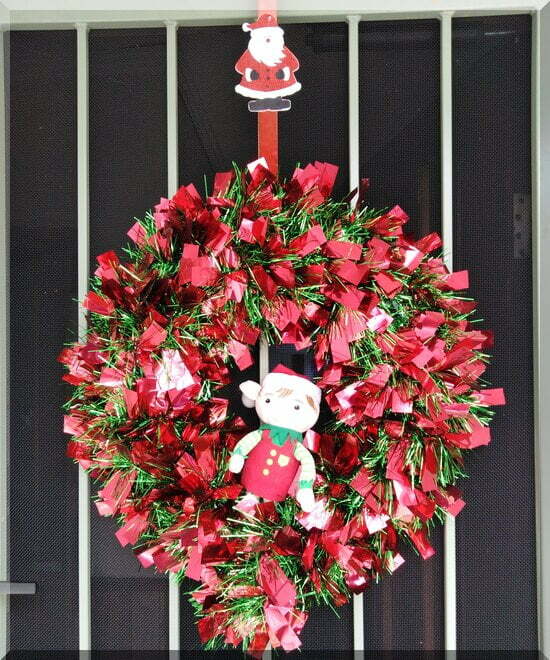 Tinkles-wreath-front-door_550 | love santa a Christmas elf sitting in a sparkling red and green wreath hanging from a Santa hook on a green door