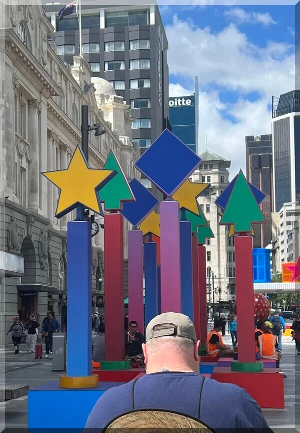 Christmas_stars_auckland_nov2023-600 | love santa With tall city buildings n the background, man views colourful pillars on which green and yellow Christmas tree and star shapes sit